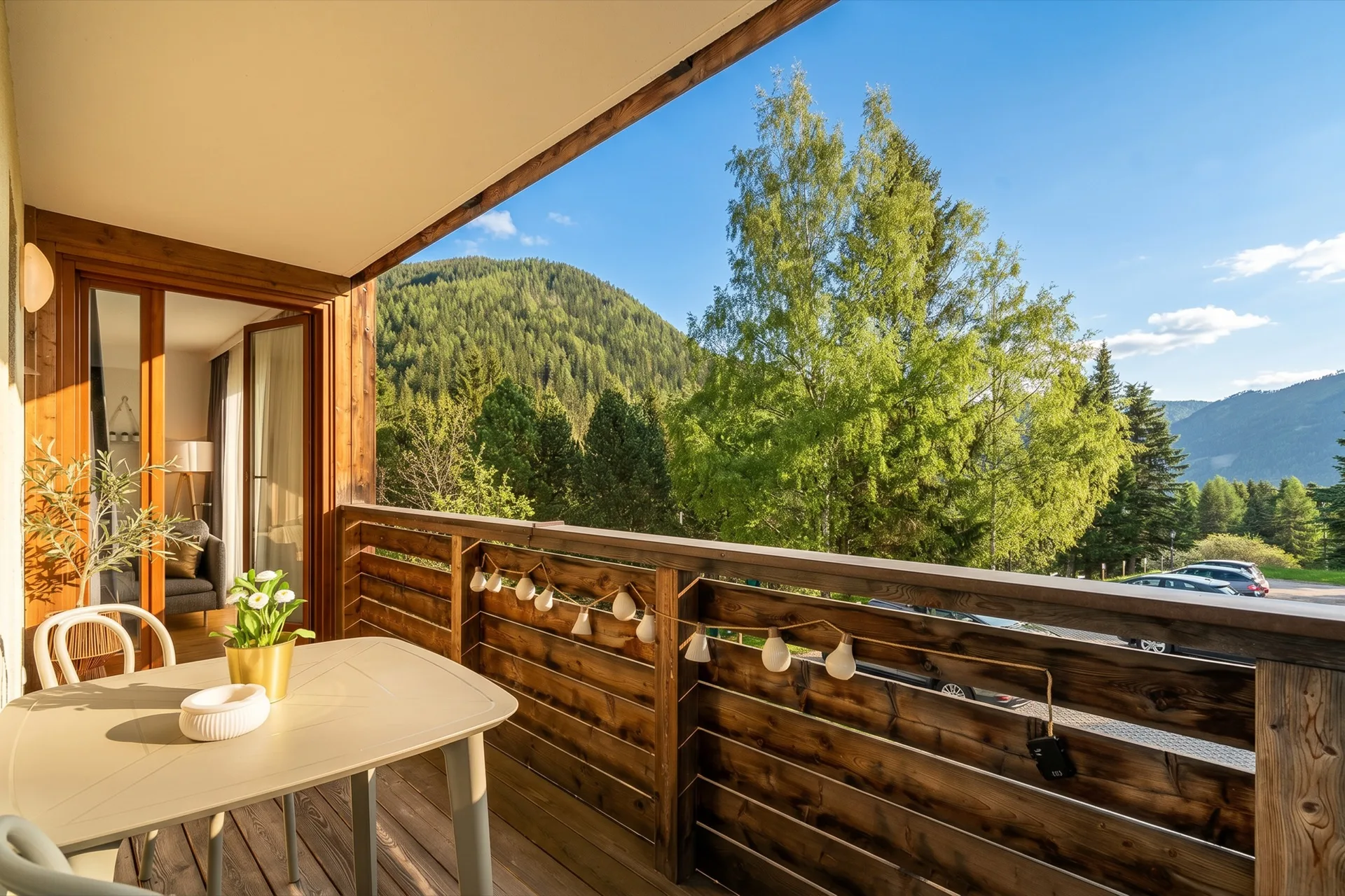 Holzbalkon mit weißem Bistrotisch, Lichterkette und warmem Abendlicht, Blick in das gemütliche Wohnzimmer und auf grüne Berglandschaft beim Alpstay Chalet Nr.1 Bad Kleinkirchheim – Balkonfotografie Ferienwohnung