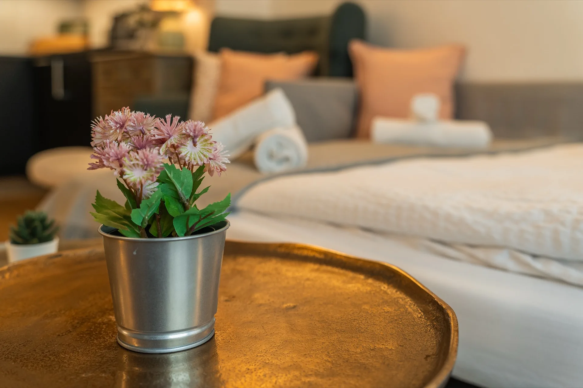 Zartrosa Blumenstrauß in Metalltopf auf goldenem Tablett mit warmem Schlafzimmer-Bokeh im Hintergrund, Alpstay Chalet Nr.2 Bad Kleinkirchheim – Atmosphäre-Detail-Fotografie Ferienwohnung Kärnten
