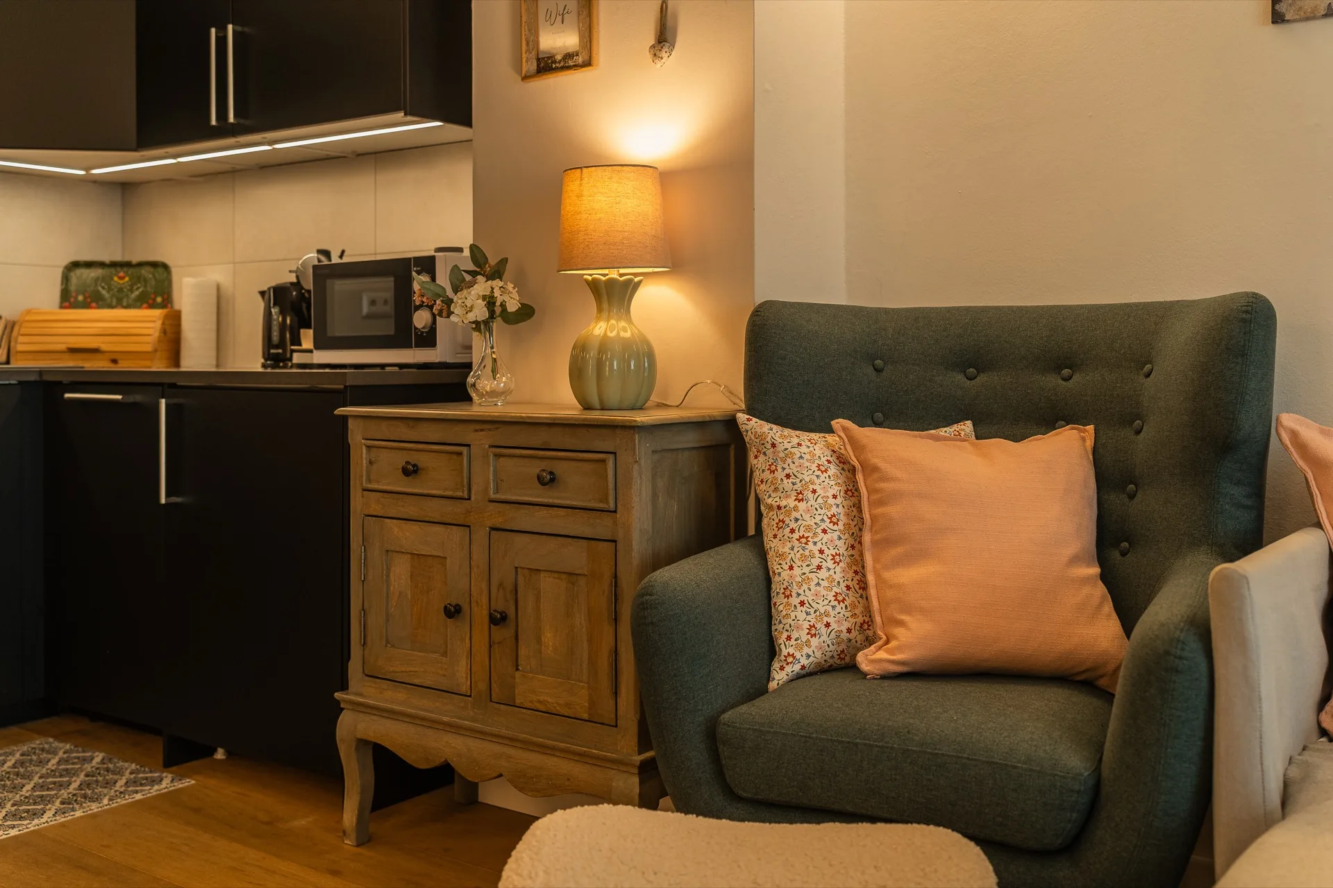 Gemütlicher grüner Ohrensessel mit rosafarbenem Kissen, warmer Tischlampe auf antikem Sideboard und sichtbarer moderner Küche im Hintergrund, Alpstay Chalet Nr.2 Bad Kleinkirchheim – Wohnatmosphäre-Fotografie Ferienwohnung Kärnten