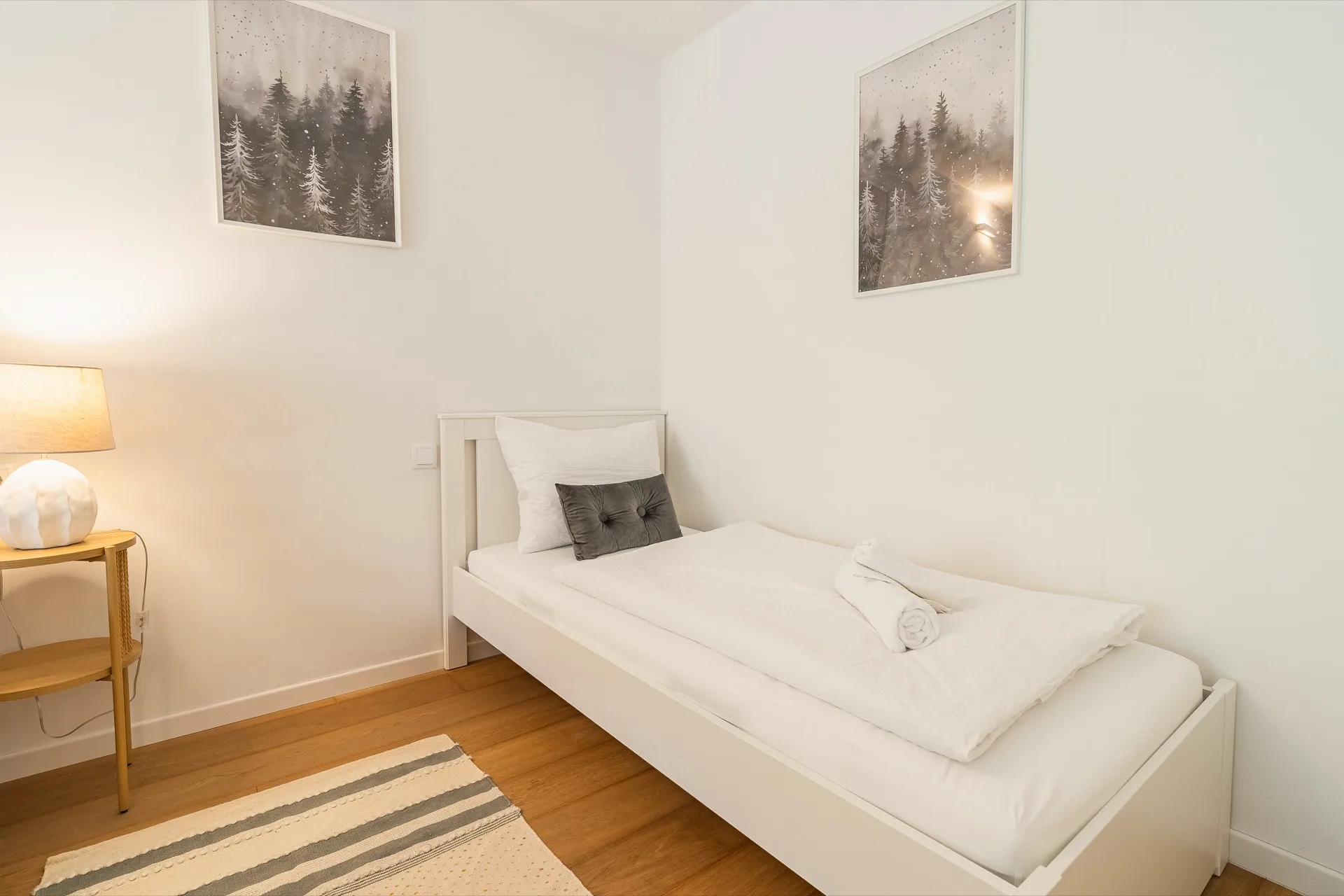 Helles Einzelzimmer mit weißem Bett, Nachttischlampe, Parkett und zwei stimmungsvollen Waldmotiv-Kunstdrucken im Alpstay Chalet Nr.2 Bad Kleinkirchheim – Schlafzimmerfotografie Ferienwohnung Kärnten